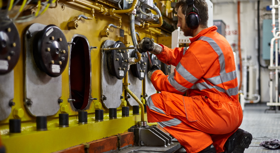 Engineer wearing high-vis flame retardant boilersuit kneels on floor to inspect engine pistons.